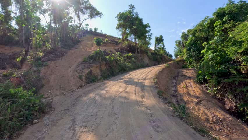Vehicle travels a sunlit, rural dirt road through lush, hilly landscape with steady forward motion