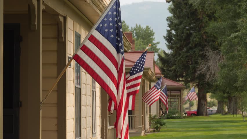 Historic homes adorned with rows of American flags on Memorial Day. Vibrant reds, whites, and blues contrast lush greenery in a nostalgic small-town USA scene. Bright daylight enhances charm.