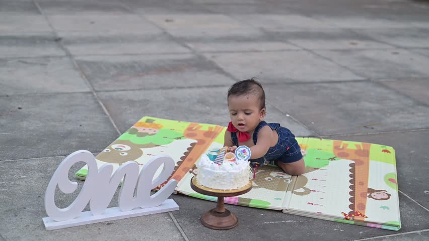 A cute baby celebrating a first birthday with a cake smash during an outdoor photoshoot. The child is sitting on a colorful play mat next to a decorated birthday cake and a “One” sign, wearing a cute 