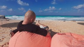 Man sits on bean bag chair at sandy beach reading a book opposite turquoise sea waves under a deep blue sky with fluffy clouds. Relaxation, vacation mood, leisure time at tropical seaside destination - Powered by Shutterstock - Get 15% off with code: PIKWIZARD15