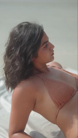 Vertical close-up of young woman in bikini lying on white fabric at beach, wind blowing hair.
