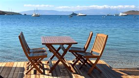 Seaside dining deck with wooden tables and chairs overlooking a beautiful turquoise bay, sailboats, and distant hills under a clear blue sky. - Powered by Shutterstock - Get 15% off with code: PIKWIZARD15