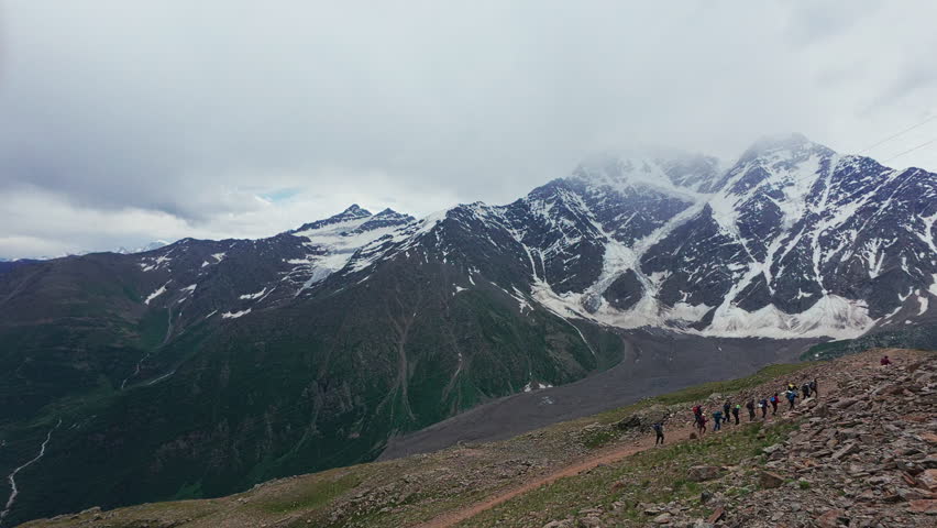 A group of hikers trekking along rocky mountain trail with snow-covered peaks rising in the background under cloudy skies. Adventure, exploration, connection with nature in high-altitude environment