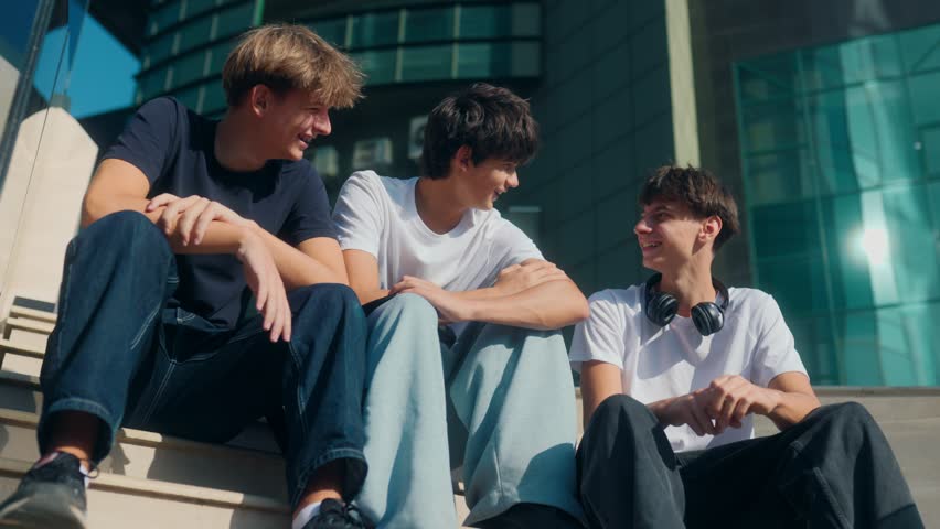 Three teenagers sit on stairs laughing and enjoying conversation. With headphones, jeans, and casual tees, they reflect modern youth, friendship, and authentic vibes.
