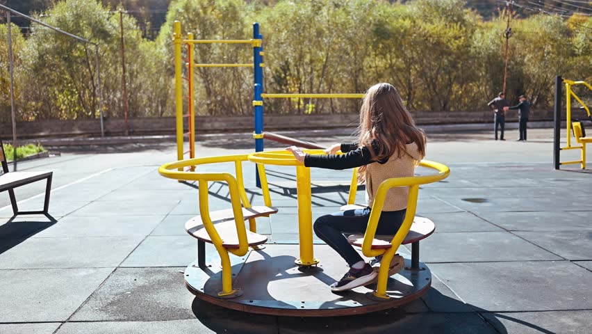 A girl sits on a yellow and black merry go round. The merry go round is in a park with a few benches and trees