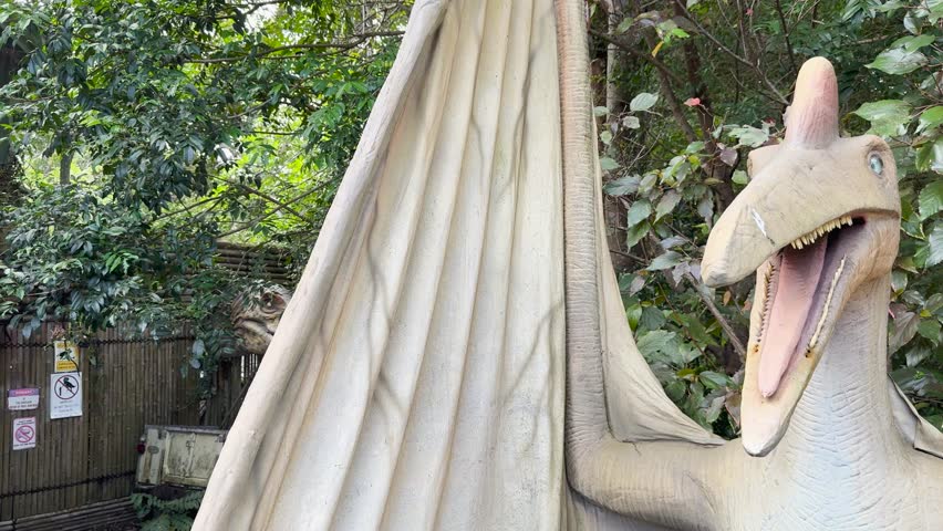 Animatronic pterosaur turns head, opens mouth in bright, leafy outdoor setting, mid-range shot