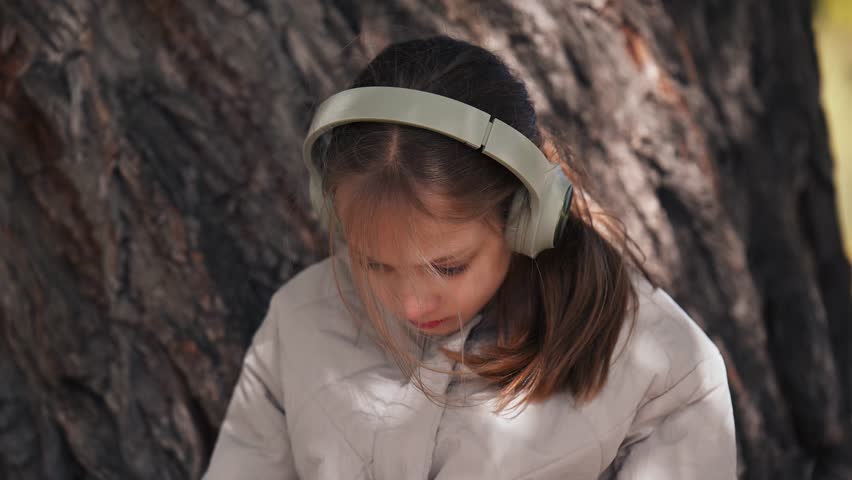 A girl wearing headphones is sitting under a tree. She is wearing a tan jacket and has her hair in a ponytail