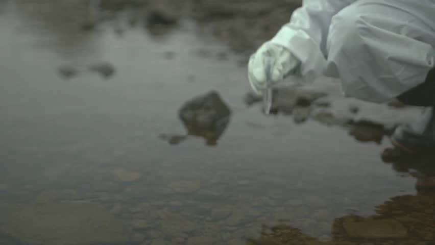 Slow-motion close-up of an ecologist in protective coveralls and gloves taking a water sample from a polluted river using a test tube during field research.

