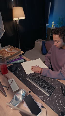 Vertical high angle view of young black male teenager having break from homework, playing video games on computer using console and headset at table