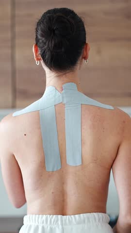 Young woman with kinesiology tape on her neck and shoulder gently massaging the area with her hand. Concept of muscle recovery, pain relief and self care using therapeutic taping techniques.