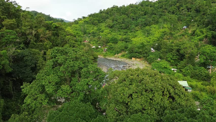 Drone view facing upstream at Tampisaw River in Barangay Napsan, Puerto Princesa. A dirt path and flagged grassy area line the right bank, with bamboo fences and small homes under lush forest.
