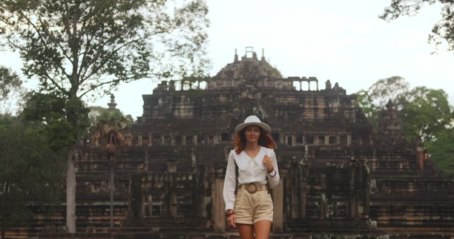 Confident female traveler in white outfit and hat walks towards the camera with Baphuon Temple in the background. High quality stock footage for travel promos, lifestyle ads, or cultural documentaries