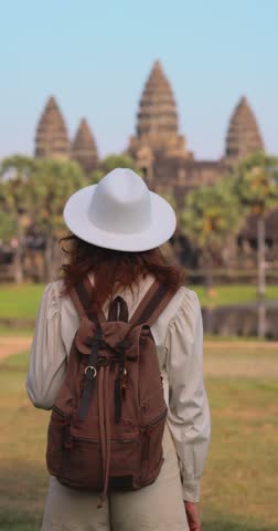 Traveler woman in white hat and backpack walks toward Angkor Wat temple in Cambodia. High quality lifestyle and tourism footage, perfect for adventure, promo, travel and cultural concept videos.