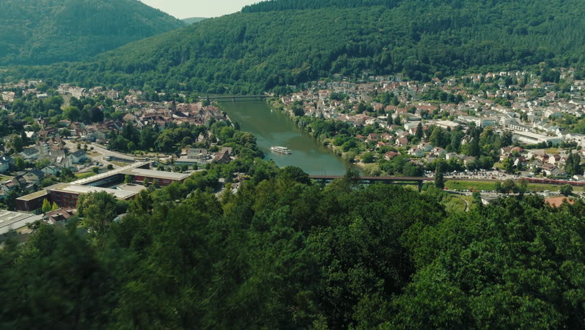 Drone shot of Neckargemünd and Kleingemünd, Germany, showing houses along the Neckar River, a train crossing the bridge, a large boat, and the surrounding forested hills on a summer day.