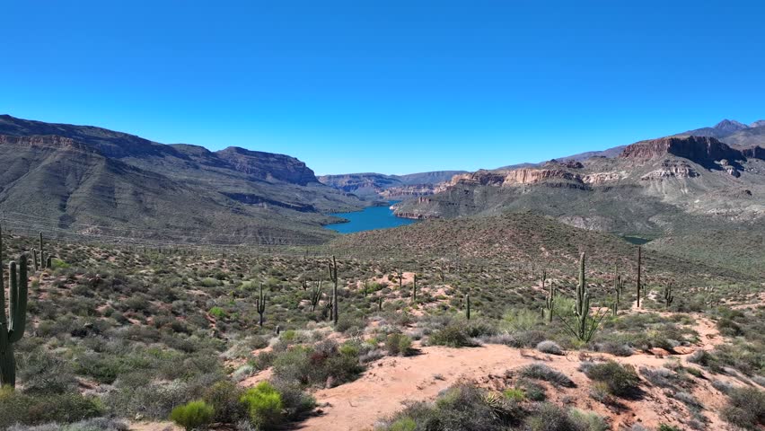 Aerial view of the Apache Trail winding through the rugged terrain towards the serene blue waters of Canyon Lake under a clear sky, Apache Junction, Arizona, United States.