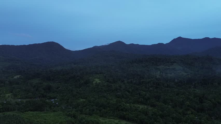 Aerial retreat reveals multilayered forested slopes with meadow clearings and hazy peaks under pre-sunset blue tones in Barangay Napsan, Puerto Princesa, Palawan, Philippines.