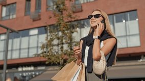 Blonde Caucasian woman with long hair wearing sunglasses while standing in middle of street. Talking on smartphone. Holding shopping bags. Enjoying conversation and carrying stylish woven handbag. - Powered by Shutterstock - Get 15% off with code: PIKWIZARD15