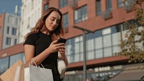 Pretty Caucasian woman with long hair holding shopping bags outdoors. Smiling and looking at smartphone screen. Browsing social media or reading messages after shopping. Modern building in background. - Powered by Shutterstock - Get 15% off with code: PIKWIZARD15