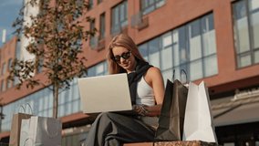 Confident Caucasian woman wearing sunglasses while sitting outdoors near modern building. Shopping bags located on bench. Holding smartphone in hand while working on laptop. Managing tasks outside. - Powered by Shutterstock - Get 15% off with code: PIKWIZARD15
