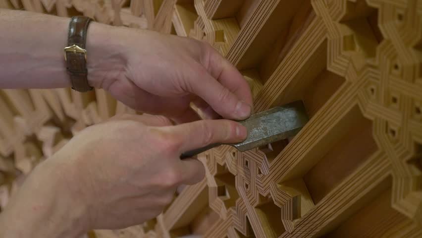 Close-up of hands restoring a Mudejar coffered ceiling, cleaning the intricate geometric patterns of carved wood with a gouge or chisel.