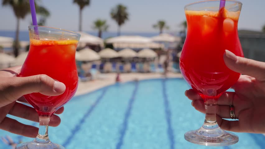  Two people toasting with refreshing fruit cocktails by a swimming pool. Concept of vacation.
