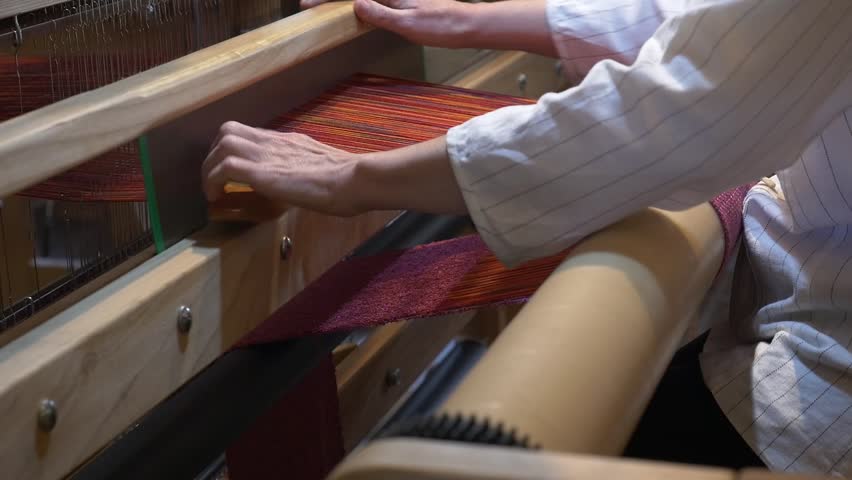 General view of the weaving process on a traditional loom. The weaver passes the shuttle between the orange warp threads, creating a raspberry-toned fabric. Handcrafted work.