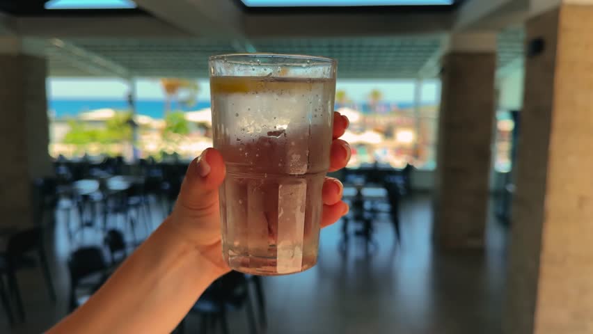 A hand holds a glass of cool sparkling water against the backdrop of the sea and palm trees. A refreshing drink that quenches thirst in the hot summer.