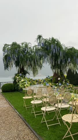 Luxury destination wedding setup on Lake Como: fresh hydrangea willow floral arch, mirrored aisle, guest chairs, soft rain, dreamy mist over Como creating poetic romantic ambience