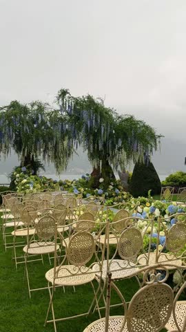 Luxury destination wedding setup on Lake Como: fresh hydrangea willow floral arch, mirrored aisle, guest chairs, soft rain, dreamy mist over Como creating poetic romantic ambience