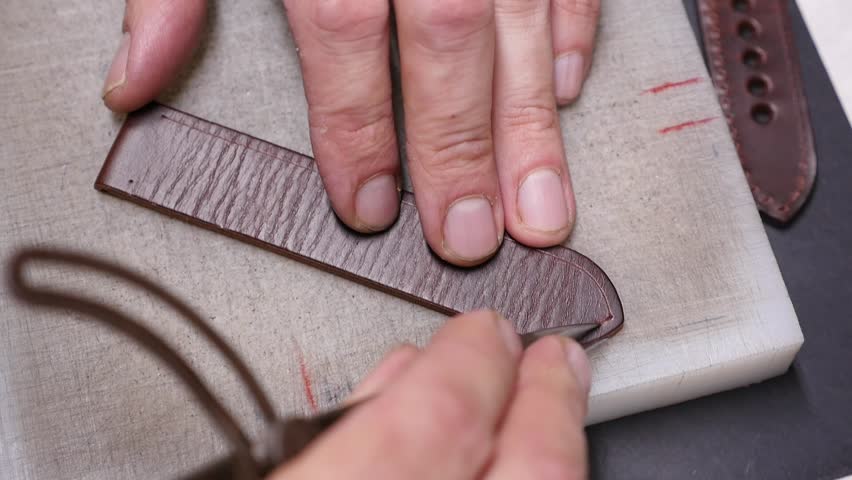 Close-up of a craftsman using a scoring tool to engrave a decorative line or stitching guide into a piece of textured brown leather, which will become a handmade watch strap.