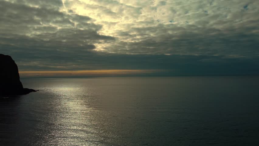 Beautiful Rising Aerial of Dark Contrasting Clouds Over Calm Ocean with Orange Sunlight on Horizon with Silhouetted Rocky Coastline.