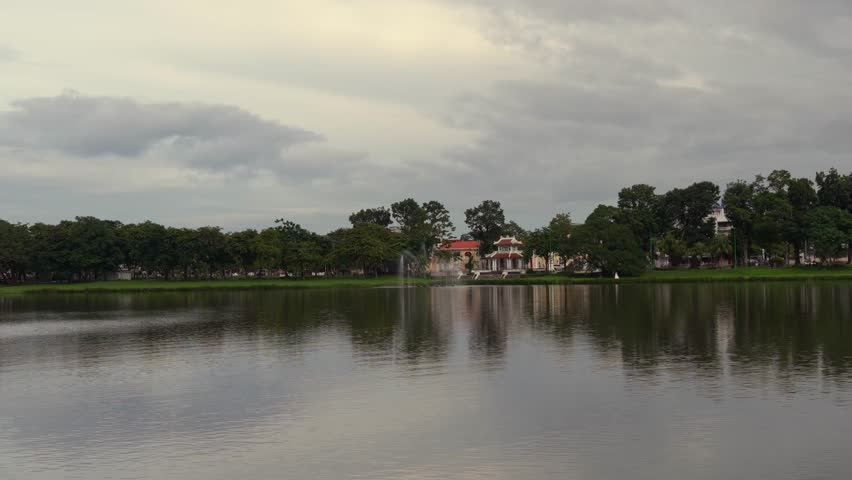 A scenic view across a calm lake in Hue, featuring a white hexagonal pavilion, a small fountain, and a dense line of trees reflecting on the water.