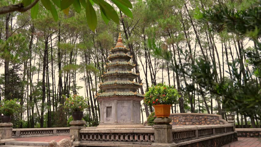an ancient, multi-tiered stone stupa (possibly at Tu Hieu Pagoda) surrounded by a dense, tall pine forest, framed by potted plants and tropical foliage.