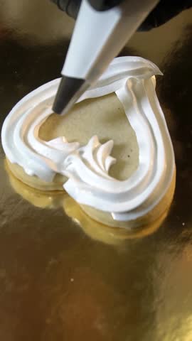 The pastry chef uses a piping bag to shape red jam onto the top of the heart-shaped cookie.