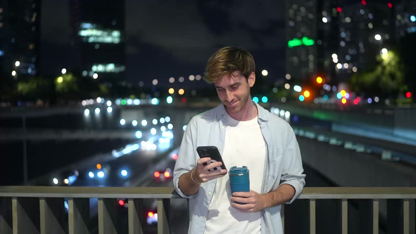 Cheerful young man standing on a city bridge at night, smiling while using his mobile phone and holding a reusable coffee cup, with blurred car lights and urban buildings in the background