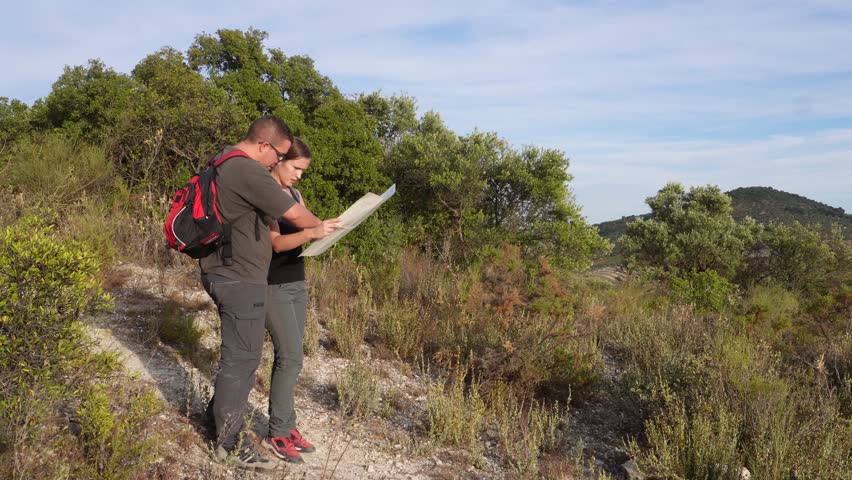 A man and woman in outdoor clothes study a paper map under warm light on a rural trail surrounded by green bushes and rocky terrain.