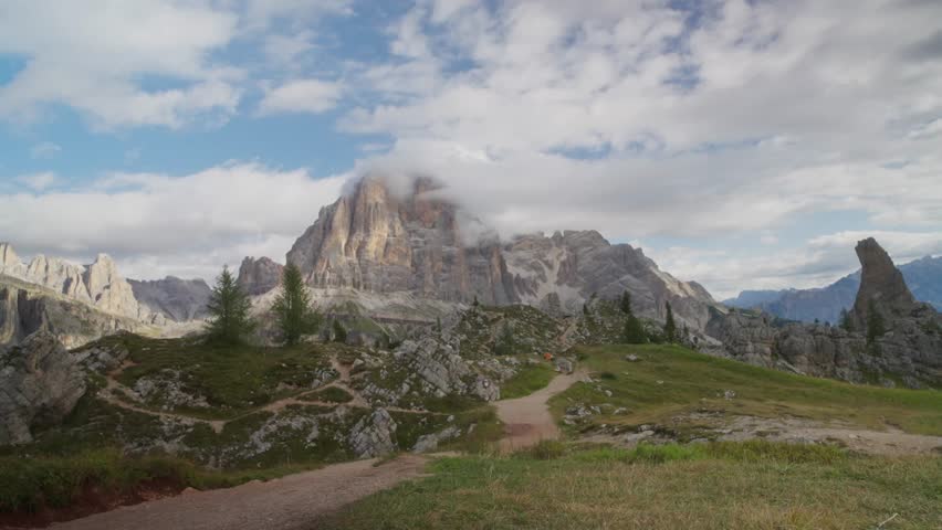 Male hiker wearing a yellow jacket and backpack walking along a rocky alpine path toward the dramatic peaks of the Dolomites in Italy.