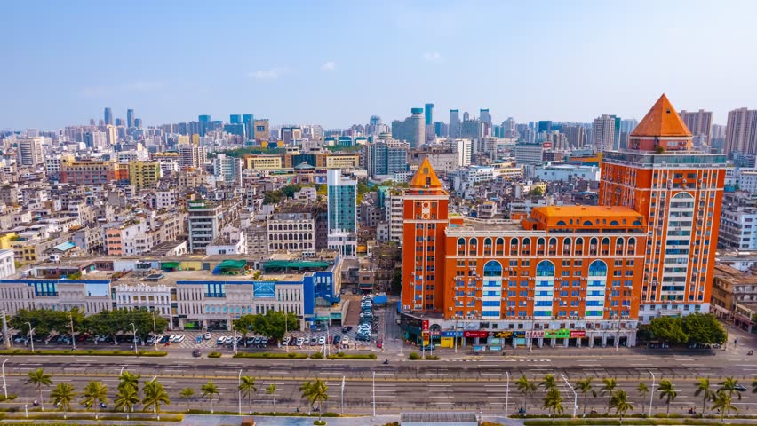 Aerial view of Haikou cityscape in Hainan, China showing modern skyscrapers, distinctive orange castle-like building, urban development and palm-lined streets under blue sky.
