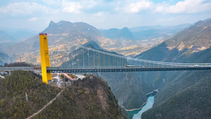 Aerial view of a massive suspension bridge spanning across a deep mountain canyon with river below, showcasing impressive engineering and natural landscape.
