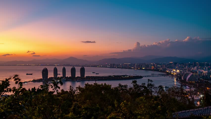 Stunning time lapse view of Sanya city skyline in Hainan, China during atmospheric transition from day to night with illuminated buildings and colorful sunset sky over water.
