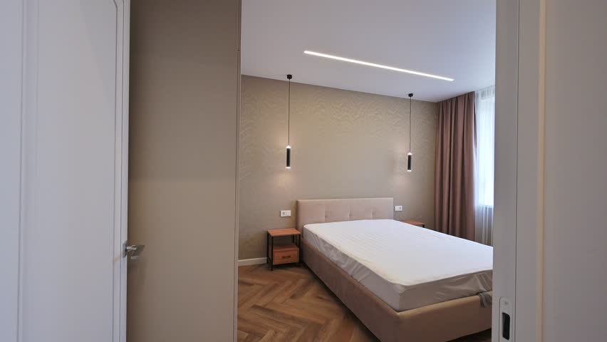 A modern bedroom features a queen-sized bed, bedside tables, pendant lights, and textured wallpaper.  Brown curtains flank a bright window