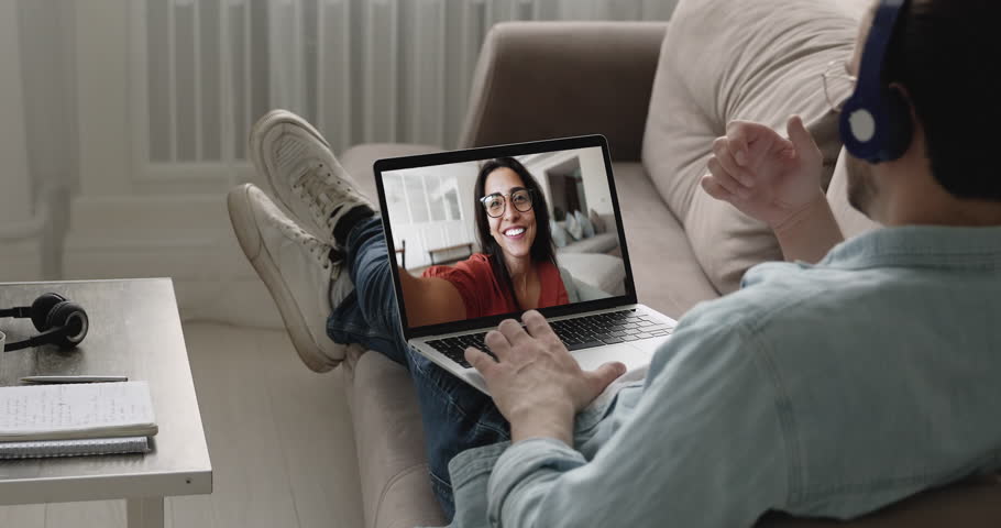Man wear headphones engaged in video call on laptop relaxing on sofa at home, screen shows smiling woman enjoy their friendly informal conversation. Long distance relationship, digital communication