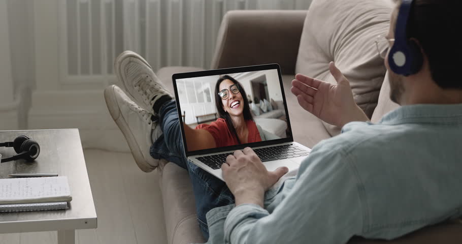 Man wear headphones engaged in video call on laptop relaxing on sofa at home, screen shows smiling woman enjoy their friendly informal conversation. Long distance relationship, digital communication