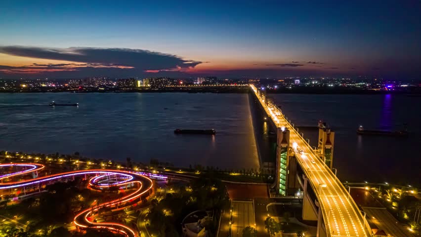 Aerial time lapse view of illuminated Jiujiang Bridge crossing Yangtze River at evening with traffic light trails and city skyline in background during twilight hour.