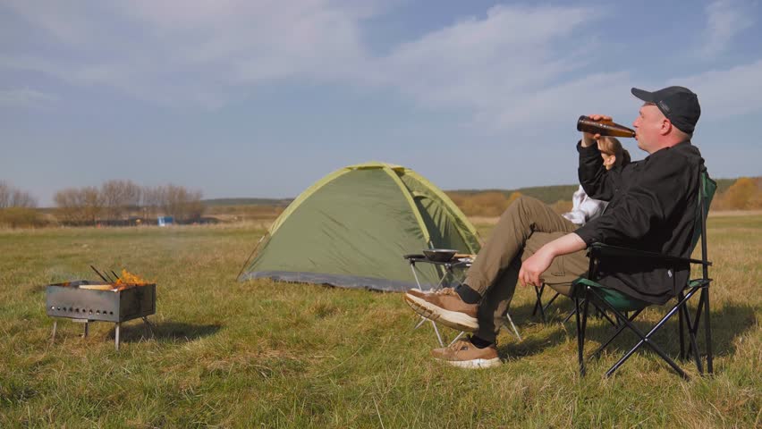 Cheerful couple enjoying a picnic lunch with beer next to their tent while camping, relaxing on foldable chairs and conversing in a scenic green meadow on a sunny spring day outdoors