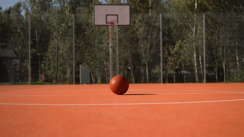 Pull in shot of basketball ball lies in center of basketball court against hoop on sunny day. Sports equipment and modern outdoor basketball court with orange surface and fencing.