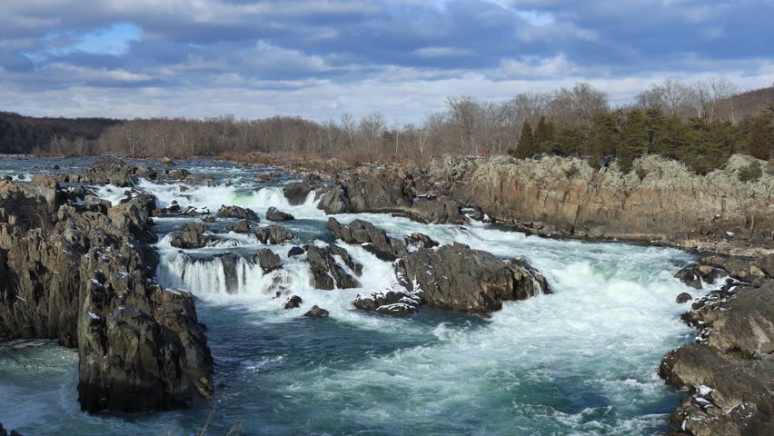 Rushing white water cascades over large, jagged rocks at Great Falls of the Potomac in Virginia. The river is framed by bare winter trees and a dramatic cloudy sky.