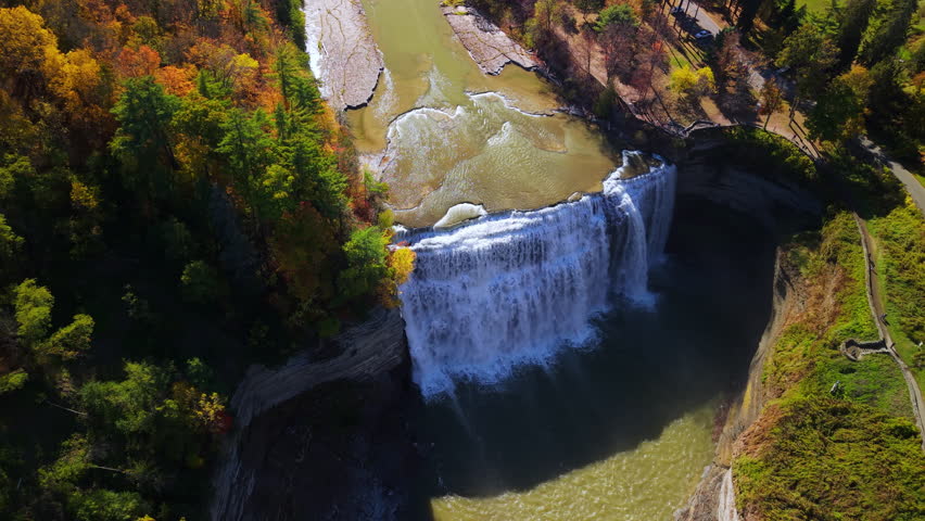 Aerial view of the majestic waterfall in Letchworth State Park, New York, surrounded by vibrant autumn foliage. The powerful cascade drops into the deep gorge below, framed by colorful forest and