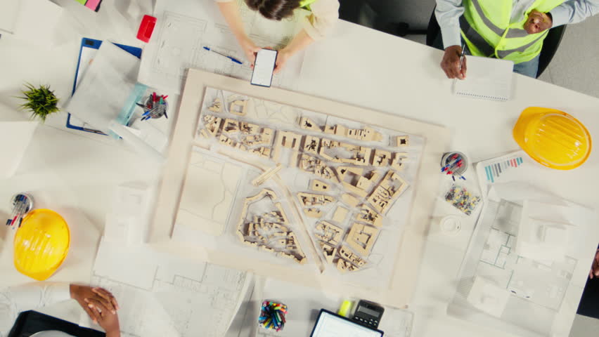 Time lapse top down view zoom out of real estate developers and architects in office reviewing blueprints and 3D models. Development team discusses urban planning and property investment. Camera A.