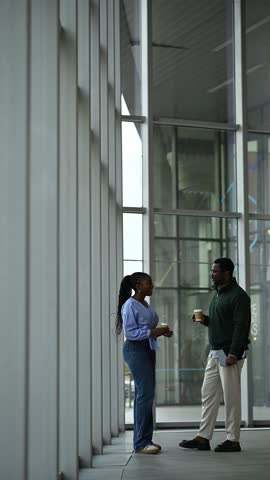 Full length shot of two young black colleagues having an informal conversation during a coffee break in a modern office building hallway